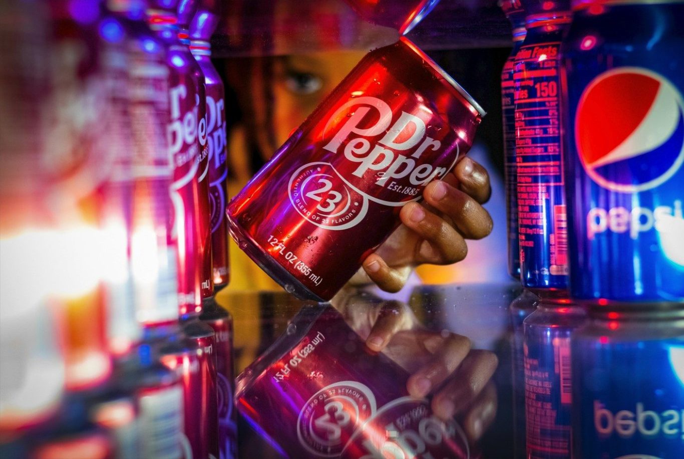 Eine Hand hält eine Dose Dr. Pepper zwischen leuchtenden Flaschen von Pepsi als Beispiel für ein gutes Brand Naming.